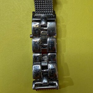 May include: A silver-toned watch band with a mesh section and rectangular links. The band is made of metal and has a polished finish. The watch band is shown against a yellow background.
