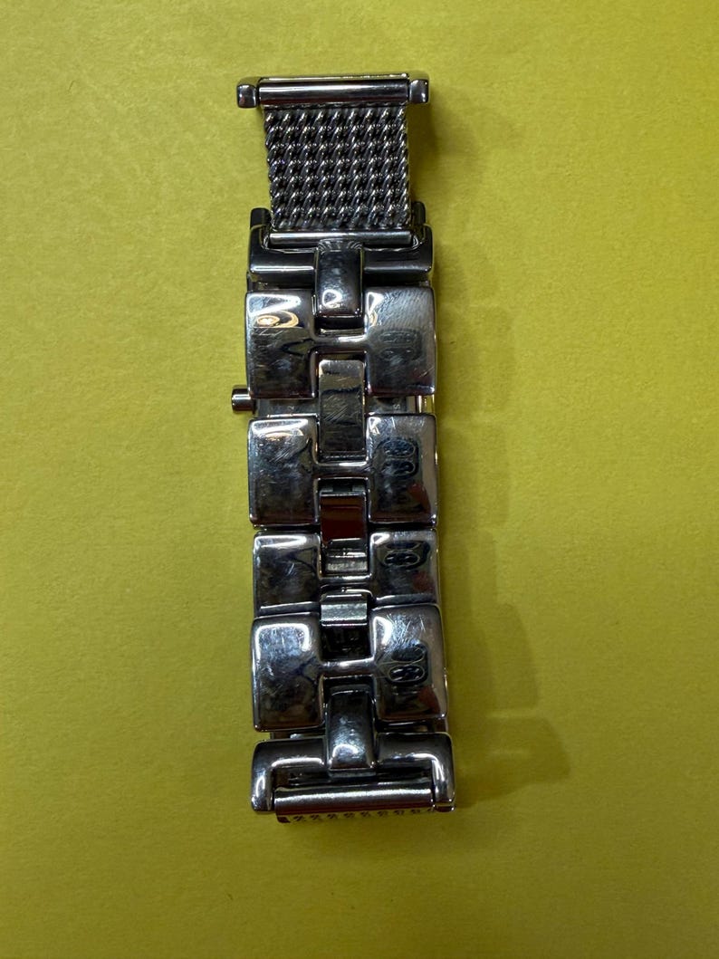 May include: A silver-toned watch band with a mesh section and rectangular links. The band is made of metal and has a polished finish. The watch band is shown against a yellow background.