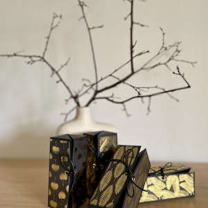 Könnte beinhalten: Schwarze und goldene Geschenkboxen mit verschiedenen Blättermustern, gebunden mit schwarzem Faden. Die Boxen stehen auf einer Holzoberfläche, im Hintergrund eine weiße Vase und Zweige. Die Boxen haben unterschiedliche Größen und Formen.