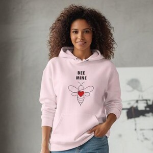 Könnte beinhalten: Hellrosa Kapuzenpullover mit dem Text "BEE MINE" über einer Bienen-Grafik mit einem roten Herz. Der Hoodie wird mit blauen Jeans und weißen Turnschuhen kombiniert. Die Person steht vor einem modernen abstrakten Gemälde.