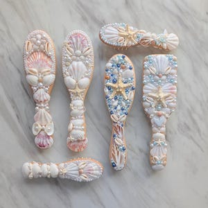 May include: Several decorative hairbrushes decorated with seashells, pearls, and starfish. The brushes have diverse designs in white, beige, and pastel shades. The handles are embellished with pearls and shells, creating a coastal theme.