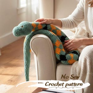 May include: A handmade crochet snake in shades of teal, orange, and black. The snake is coiled on a chair, with the text "No Sew Crochet pattern" visible at the bottom. The snake's head is detailed with small yellow eyes.