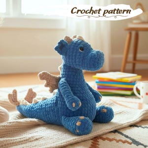 Puede incluir: Un dragón azul tejido a ganchillo con alas, cuernos y almohadillas para los pies de color beige. El dragón está sentado sobre una alfombra color crema. El texto "Crochet pattern" está encima del dragón.