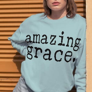 May include: A light blue sweatshirt with the words "amazing grace" in black lettering. The person is wearing sunglasses and denim shorts. The background is a textured orange wall.
