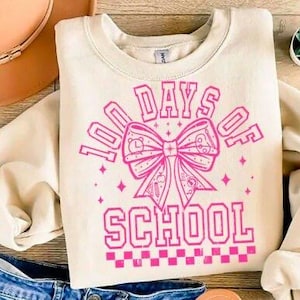 May include: Cream-colored sweatshirt with the text "100 DAYS OF SCHOOL" in bright pink. The design features a large bow with school-themed icons. The sweatshirt is laid on a wooden surface with a hat and jeans.