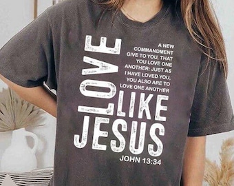 Può includere: T-shirt grigio scuro con le parole "LOVE LIKE JESUS" a grandi lettere bianche. La maglietta presenta anche un versetto biblico di Giovanni 13:34. Una maglietta casual e confortevole.