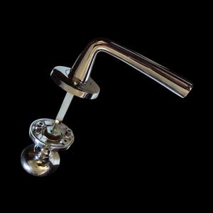 Op de afbeelding: Een glanzende chromen deurklink tegen een zwarte achtergrond. De klink heeft een gebogen hendelontwerp en een ronde basis. De set bevat een spil en een bolvormige knop, die allemaal licht reflecteren.