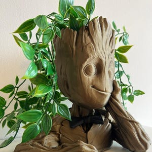 Puede incluir: Una maceta Groot marrón impresa en 3D con una pajarita negra, sosteniendo su barbilla. La maceta está llena de plantas verdes con hojas. El artículo es decorativo.