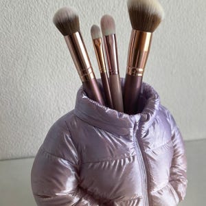 May include: A lavender puffer jacket-shaped makeup brush holder, holding several makeup brushes. The brushes have brown and gold handles and soft bristles. The jacket has a shiny, quilted texture, and is a decorative item.