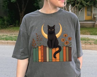 May include: Dark gray t-shirt featuring a black cat sitting on a stack of books. The cat is in front of a crescent moon and flowers. The books are in shades of green, orange, and brown.
