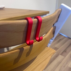 May include: Two bright red hooks are attached to the back of a wooden high chair. The chair has a natural wood finish and a white support structure. The hooks are designed to hang items.