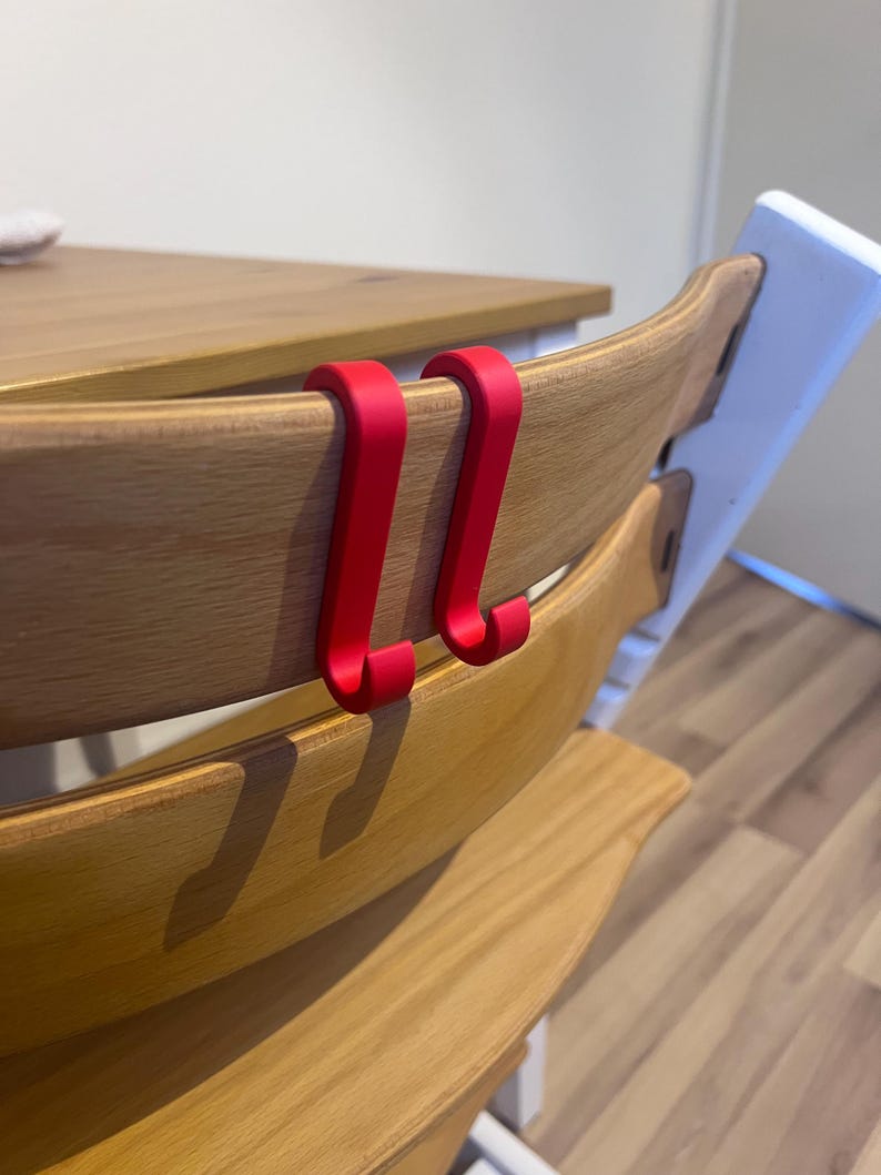 May include: Two bright red hooks are attached to the back of a wooden high chair. The chair has a natural wood finish and a white support structure. The hooks are designed to hang items.