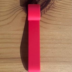 May include: A bright red, rectangular object with rounded edges, possibly a bracelet or a similar accessory, is positioned vertically against a light brown wooden surface. The object casts a shadow to its left, enhancing its three-dimensional appearance.