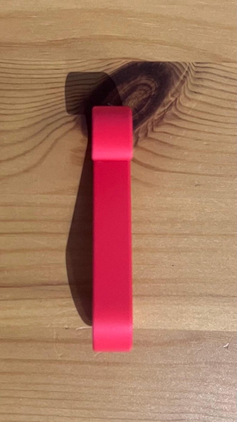 May include: A bright red, rectangular object with rounded edges, possibly a bracelet or a similar accessory, is positioned vertically against a light brown wooden surface. The object casts a shadow to its left, enhancing its three-dimensional appearance.