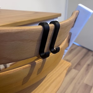 May include: Two black metal hooks are attached to the back of a wooden high chair. The high chair has a light wood finish and a white back. The hooks are designed to hang items.