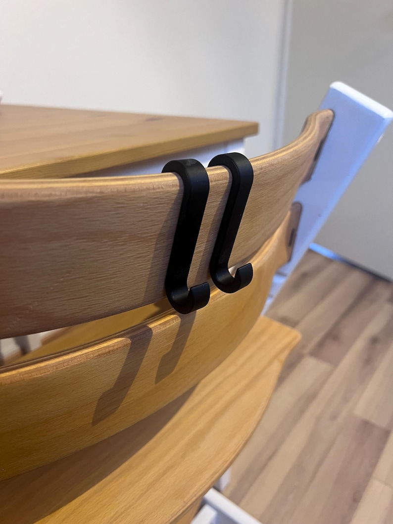 May include: Two black metal hooks are attached to the back of a wooden high chair. The high chair has a light wood finish and a white back. The hooks are designed to hang items.