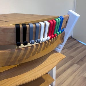 May include: A wooden high chair with a natural finish, featuring a row of colorful hooks in black, gray, white, red, green, and blue. The hooks are designed to hang items. The chair has a white frame and is set on a light wood floor.