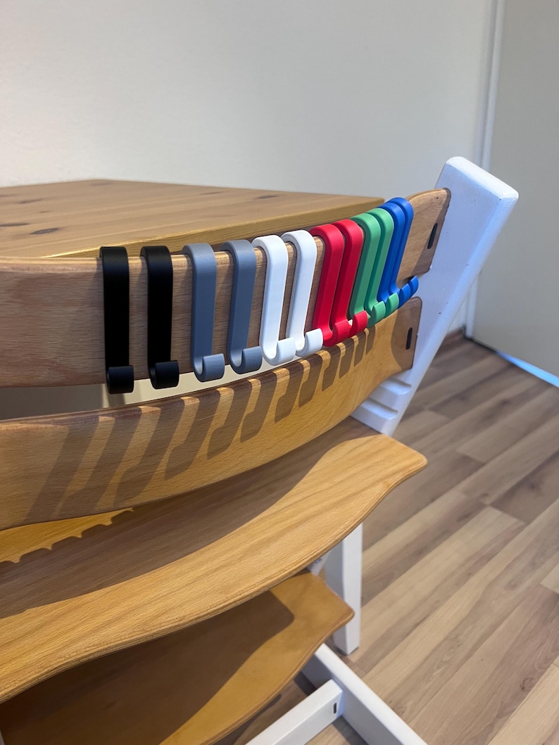 May include: A wooden high chair with a natural finish, featuring a row of colorful hooks in black, gray, white, red, green, and blue. The hooks are designed to hang items. The chair has a white frame and is set on a light wood floor.