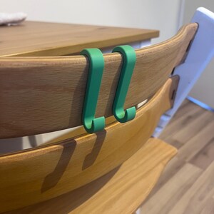 May include: Two green, curved hooks are attached to the back of a wooden chair. The chair has a light wood finish. The hooks are designed to hang items. The background includes a wooden table and a white chair.