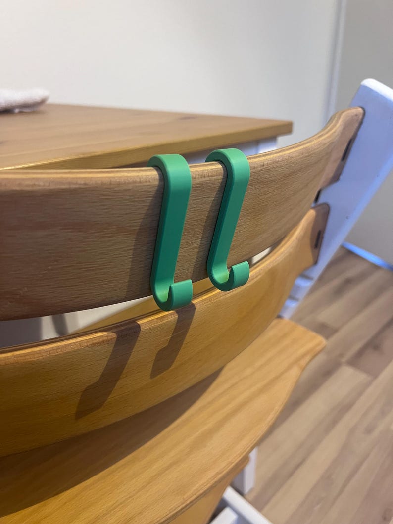 May include: Two green, curved hooks are attached to the back of a wooden chair. The chair has a light wood finish. The hooks are designed to hang items. The background includes a wooden table and a white chair.