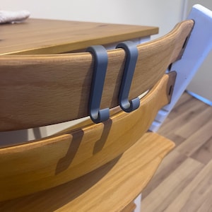May include: Two gray, curved hooks attached to the wooden back of a high chair. The high chair has a natural wood finish and is positioned near a wooden table. The hooks appear to be made of a durable plastic or metal.