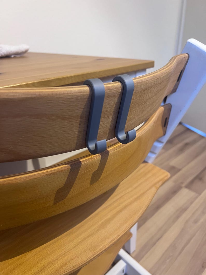 May include: Two gray, curved hooks attached to the wooden back of a high chair. The high chair has a natural wood finish and is positioned near a wooden table. The hooks appear to be made of a durable plastic or metal.