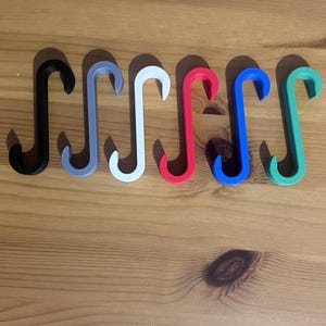 May include: A collection of seven S-shaped hooks in various colors, including black, gray, white, red, blue, and green. The hooks are arranged in a row on a wooden surface. These hooks are likely used for hanging items.