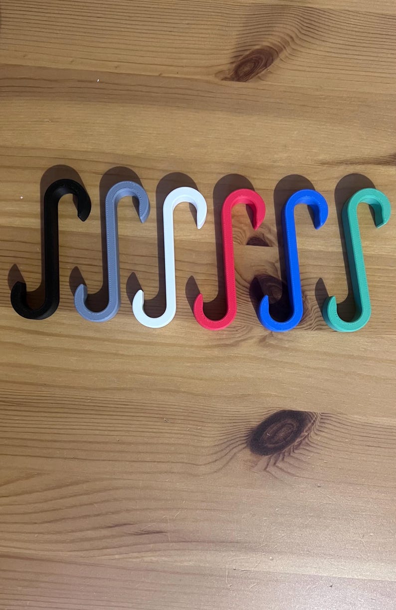 May include: A collection of seven S-shaped hooks in various colors, including black, gray, white, red, blue, and green. The hooks are arranged in a row on a wooden surface. These hooks are likely used for hanging items.