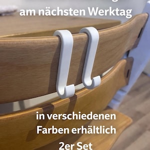 May include: Two white, curved hooks designed to attach to a wooden chair. The hooks are shown attached to the chair's backrest. Text in the image says "Versand in der Regel am n&auml;chsten Werktag" and "in verschiedenen Farben erh&auml;ltlich 2er Set."