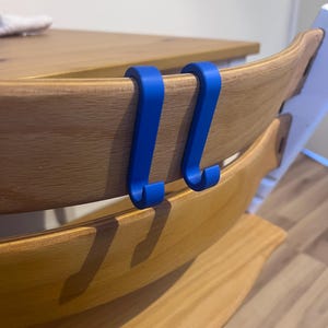 May include: Two bright blue hooks are attached to the back of a wooden high chair. The hooks are designed to hang items. The high chair has a natural wood finish.