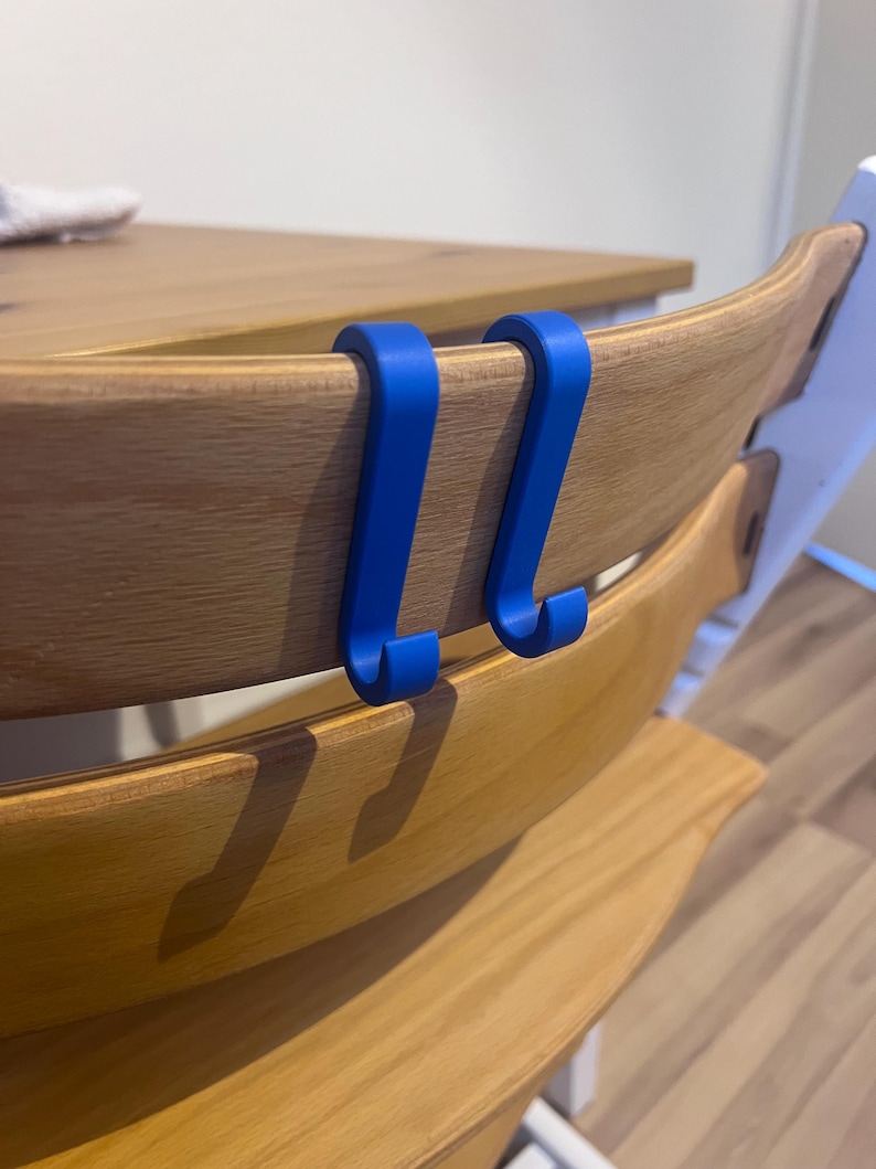 May include: Two bright blue hooks are attached to the back of a wooden high chair. The hooks are designed to hang items. The high chair has a natural wood finish.