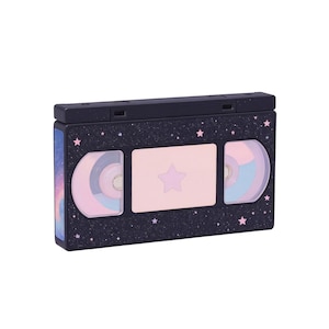 May include: A black cosmetic case shaped like a VHS tape, featuring a starry night design with pink stars. The case has a pink star in the center and two circular areas with pastel colors. The side has a blue and purple galaxy design.