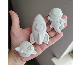 May include: A collection of white space-themed figurines, including a rocket ship, an astronaut, and a planet with a ring. The figurines are small and appear to be made of a solid material. The astronaut has a star on its chest.