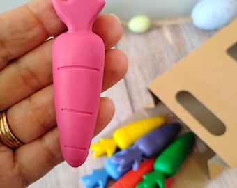 May include: A close-up of a pink carrot-shaped crayon held in a hand, with a gold ring on the finger. Several colorful carrot crayons are visible in a brown cardboard box. The crayons are in shades of blue, orange, red, green, purple, and yellow.