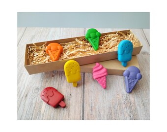 May include: A collection of colorful ice cream-shaped crayons in various colors, including orange, green, blue, yellow, pink, purple, and red. The crayons are arranged on a wooden surface, with some inside a brown box filled with shredded paper.