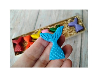 May include: A hand holds a bright blue mermaid tail-shaped crayon, with a box of colorful mermaid tail crayons in the background. The box contains red, yellow, green, pink, and purple crayons. The crayons are on a bed of brown paper.