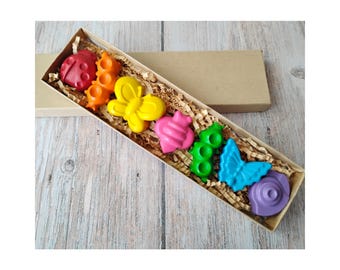 May include: A set of colorful, shaped crayons in a rectangular box. The crayons are in the shapes of a ladybug, butterfly, and snail. The crayons are red, orange, yellow, pink, green, blue, and purple. The box is filled with shredded paper.