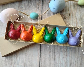 May include: A box of six colorful bunny-shaped crayons in red, orange, yellow, green, blue, and purple. The crayons are arranged in a cardboard box with straw, and are set against a wooden background with decorative Easter eggs.