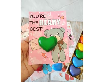 May include: A pink greeting card with the text "YOU'RE the BEARY BEST!" features a cartoon bear and a green heart-shaped object. Other cards and heart-shaped crayons are visible in the background.