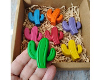 May include: A set of six cactus-shaped crayons in a variety of colors: blue, orange, purple, pink, red, green, and yellow. The crayons are displayed in a brown cardboard box filled with shredded paper.