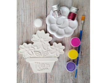 May include: A craft kit with a flower-shaped ceramic piece, paint, glitter, and a paintbrush. The ceramic piece is white and has a flower design with the word "Mom" on the pot. The paint colors are pink, yellow, and purple.