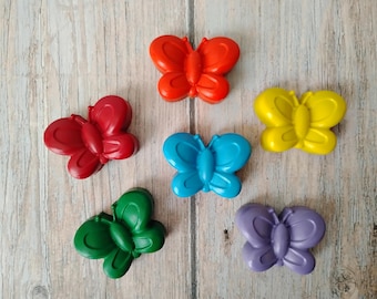 May include: Six colorful butterfly-shaped objects in red, yellow, blue, green, and purple are arranged on a light wood-grain surface. The butterflies are made of a solid material and have a smooth, glossy finish.