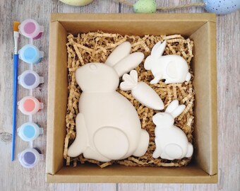May include: A craft kit featuring a cardboard box filled with white ceramic Easter bunnies and a carrot, nestled in brown paper. Paint pots in various colors and a paintbrush are included, perfect for a fun DIY project.