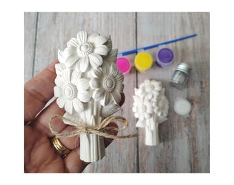 May include: A white plaster flower bouquet, tied with twine, held in a hand. Additional bouquet, paint pots in pink, yellow, and purple, a paintbrush, and glitter are on a wooden surface. Craft supplies for painting.