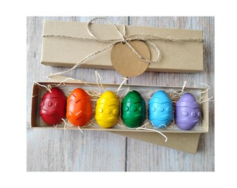 May include: A set of six colorful Easter egg-shaped crayons in a cardboard box. The crayons are red, orange, yellow, green, blue, and purple. The box is tied with twine and includes a blank tag. The crayons are nestled in straw.