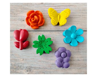 May include: A collection of colorful, decorative floral and butterfly-shaped embellishments. The assortment includes a bright orange rose, a yellow butterfly, a red tulip, a green flower, a blue flower, and a purple flower. These are likely used for crafts or decorations.