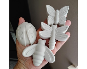 May include: Four white insect-shaped decorative items: a ladybug, a butterfly, a dragonfly, and a bee. The ladybug is oval with circular details. The butterfly has detailed wings. The dragonfly and bee have elongated bodies and wings.