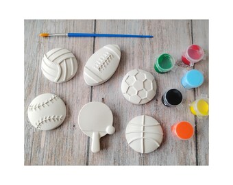 May include: A collection of white plaster sports-themed shapes, including a volleyball, football, soccer ball, baseball, ping pong paddle, and basketball. Paint pots in various colors and a blue paintbrush are also present.