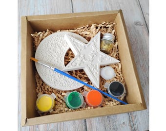 May include: A craft kit in a cardboard box, featuring a white star and crescent moon, paint pots in yellow, green, and orange, a paintbrush, and a small jar of gold glitter. The kit is designed for creative projects.