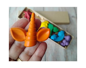 May include: An orange unicorn horn crayon is held in a hand, with a gold ring on the finger. A box of rainbow-colored crayons is in the background. The crayons are shaped like a horn, flowers, and other shapes.